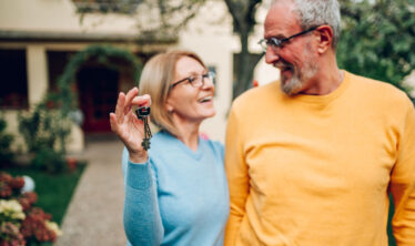 older-couple-holding-keys-standing-in-front-of-home